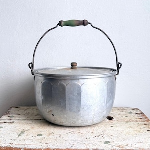 May include: A round aluminum pot with a lid and a metal bail handle featuring a green wooden grip. The pot has a scalloped, vertical panel design around its exterior body and a small, round wooden knob centered on the top of the lid.