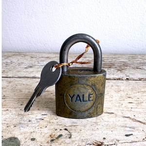 May include: Vintage Yale padlock with a key. The padlock is made of brass and has a dark, aged appearance. The key is silver and rests next to the lock. The word "YALE" is embossed on the front of the lock. A small piece of twine is attached to the lock's handle.