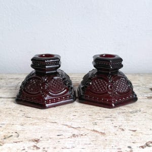 May include: A pair of deep red glass candle holders featuring a square base and a raised geometric pattern. Each piece has a central circular motif surrounded by a diamond-shaped grid texture, with a stepped neck leading to a circular opening.