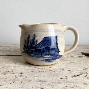 May include: A cream-colored ceramic creamer featuring a hand-painted blue illustration of a rustic barn surrounded by trees. The pitcher has a rounded body, a small pouring spout, and a curved handle, with a smooth, glossy finish throughout.