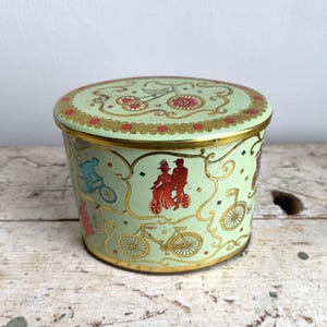 May include: A vintage, round tin with a lid, featuring a light green background and gold trim. The tin is decorated with bicycle illustrations in red, blue, and gold. The lid has a repeating pattern of red and gold accents.