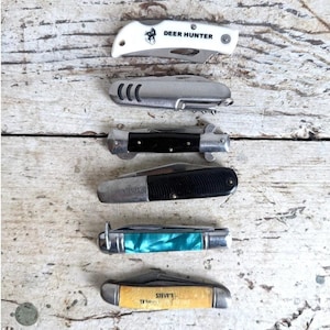以下が含まれることがあります： さまざまなハンドルデザインとブレードスタイルの6つのポケットナイフのコレクション。ナイフには、白い「Deer Hunter」ナイフ、シルバーのナイフ、黒とシルバーのナイフ、黒のナイフ、ターコイズのナイフ、「STEVE'S TRAVEL」がプリントされたナイフが含まれています。