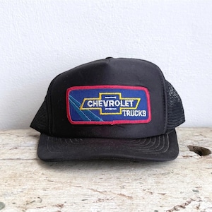 May include: A black trucker cap featuring a rectangular patch on the front. The patch has a blue background with a yellow Chevrolet logo and the text CHEVROLET TRUCKS in white, accented by a diagonal teal stripe and a red border.