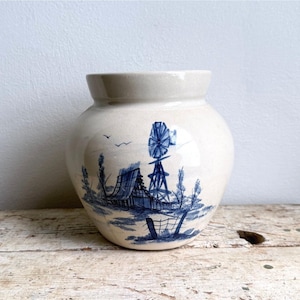 May include: A cream-colored ceramic vase with a rounded body and a wide opening. The exterior features a blue hand-painted illustration of a rural landscape, including a windmill, a small house, trees, and birds flying in the sky.