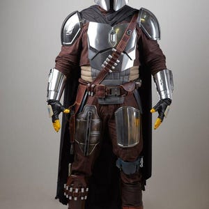 May include: Full-body shot of a detailed costume inspired by a science fiction character. The costume features a silver helmet, chest armor, and arm guards. The outfit includes brown pants, boots, and a black cape, with yellow gloves and a utility belt.