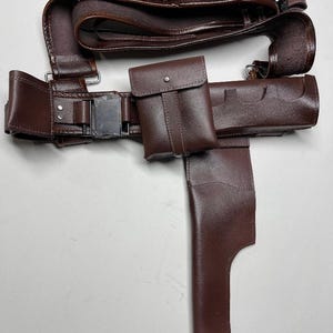 May include: A brown leather utility belt with multiple pouches, a holster, and cartridge holders. The belt features a silver buckle and is designed for carrying various items. The leather has a smooth finish.
