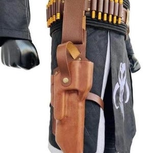 Op de afbeelding: Een bruin leren holster met bijpassende riem, met een reeks kleine cilinders met gouden uiteinden. Het holster is bevestigd aan een zwart kledingstuk met witte rand en een gestileerd embleem. De outfit is geaccessoireerd met een zwarte handschoen.