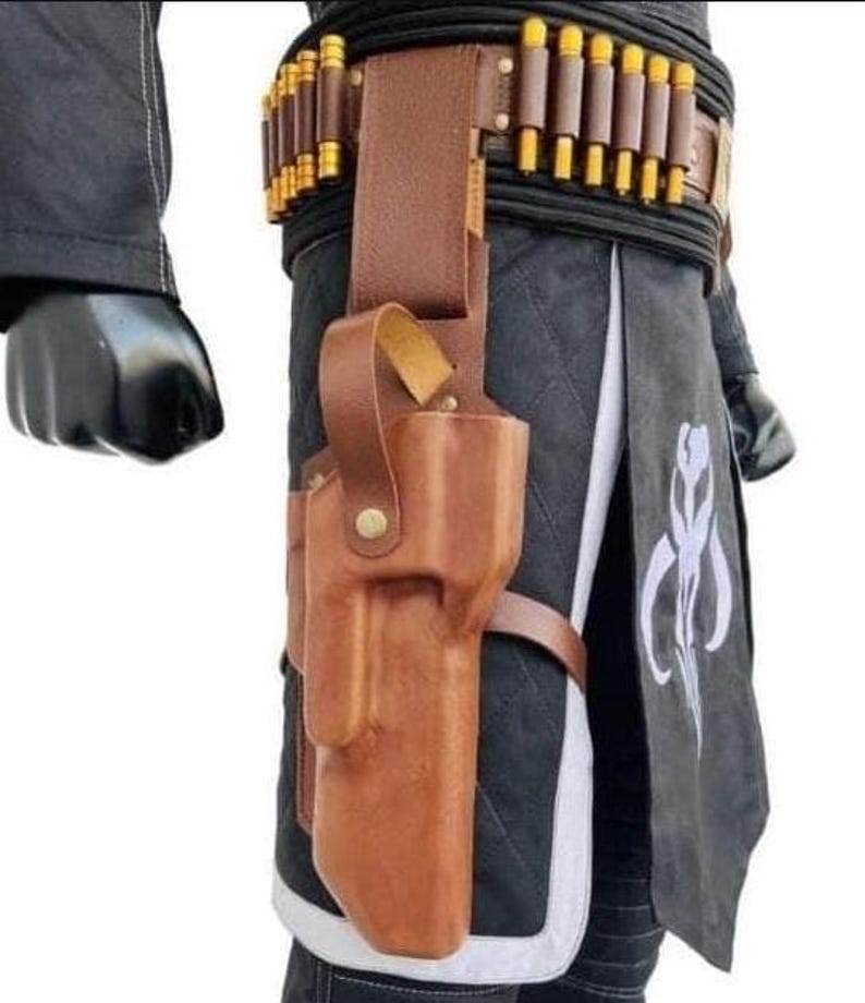 Op de afbeelding: Een bruin leren holster met bijpassende riem, met een reeks kleine cilinders met gouden uiteinden. Het holster is bevestigd aan een zwart kledingstuk met witte rand en een gestileerd embleem. De outfit is geaccessoireerd met een zwarte handschoen.