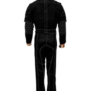 K&ouml;nnte beinhalten: Schwarzer Overall mit wei&szlig;en Steppn&auml;hten. Das Kleidungsst&uuml;ck hat einen hohen Kragen, einen G&uuml;rtel in der Taille und geometrische Muster an den &Auml;rmeln. Der Overall ist mit sichtbaren N&auml;hten versehen.