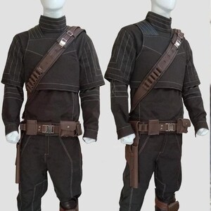 May include: Two black costumes with brown detailing are displayed on mannequins. Each outfit features a high-collared top, trousers, and a utility belt with pouches. A brown shoulder strap with ammunition holders and a holster are also included. White stitching details the seams.