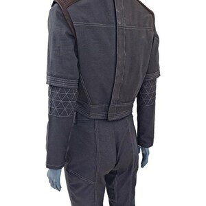 May include: Dark gray jumpsuit with brown shoulder accents and geometric stitching on the sleeves. The jumpsuit features a high collar and a fitted waist. The back of the jumpsuit has vertical paneling and visible stitching.