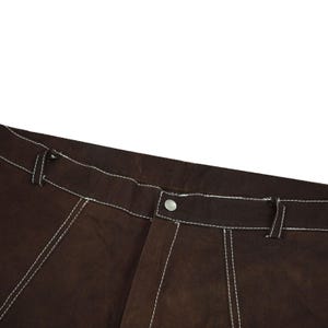 May include: Close-up of brown pants with white stitching. The pants have a button closure and a classic design. The white stitching provides a contrasting detail to the brown fabric. The image focuses on the upper portion of the pants.