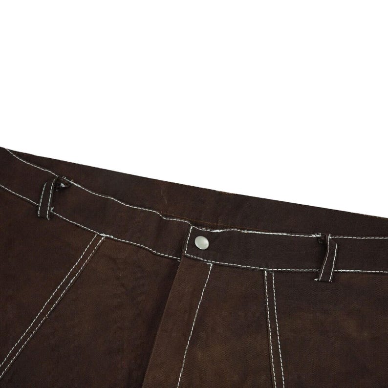 May include: Close-up of brown pants with white stitching. The pants have a button closure and a classic design. The white stitching provides a contrasting detail to the brown fabric. The image focuses on the upper portion of the pants.