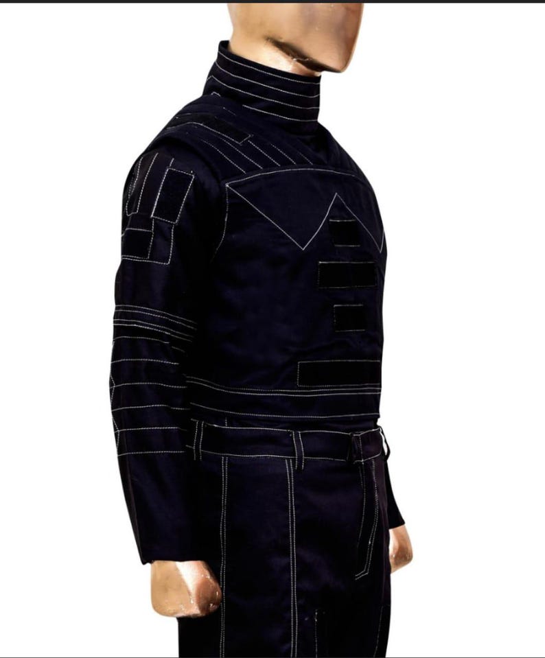 K&ouml;nnte beinhalten: Dunkelmarineblauer Overall mit wei&szlig;en Steppdetails. Der Overall hat einen hohen Kragen, lange &Auml;rmel und einen G&uuml;rtel. Das Design umfasst geometrische Formen und Stepplinien &uuml;ber Brust, Schultern und &Auml;rmeln.