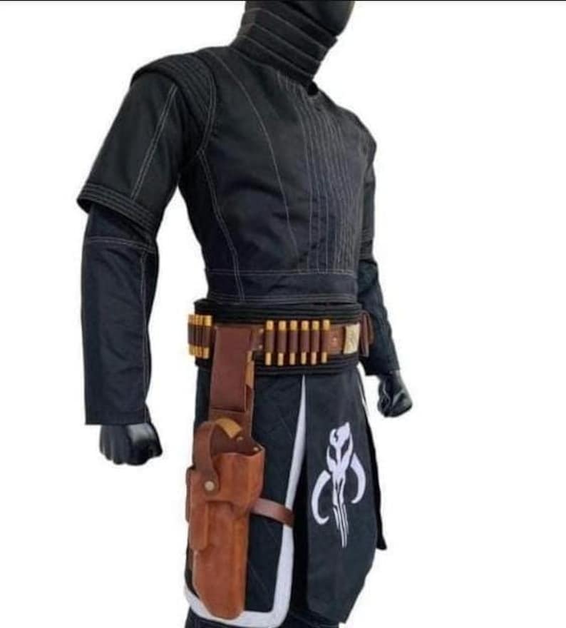 Op de afbeelding: Zwart kostuum met witte stiksels, met een hoge kraag en een gereedschapsriem met bruine accenten. Een bruin holster is aan de riem bevestigd. Het kostuum bevat een zwarte rok met een wit embleem. Dit is een cosplay-outfit.