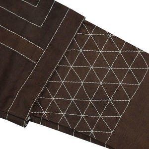 May include: Close-up of a section of brown fabric with white stitching. The design includes straight lines and a geometric pattern of triangles. The stitching is prominent against the dark background, creating a textured effect.