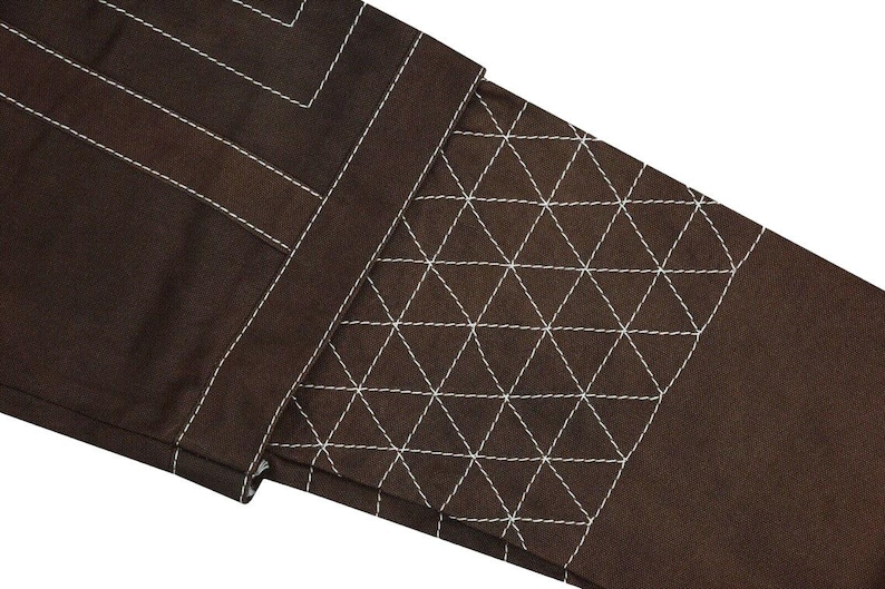 May include: Close-up of a section of brown fabric with white stitching. The design includes straight lines and a geometric pattern of triangles. The stitching is prominent against the dark background, creating a textured effect.