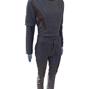 May include: A two-piece dark grey outfit on a mannequin. The top is a cropped jacket with a high collar and long sleeves. The pants are high-waisted with pockets and patches. White stitching details the seams and accents the design.