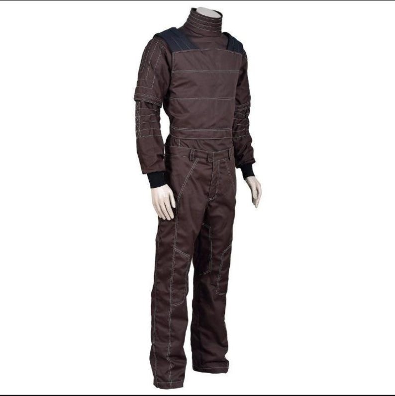 May include: Brown jumpsuit with a high collar and white stitching details. The long-sleeved top has shoulder pads and the pants have pockets. Black cuffs are visible at the wrists and ankles. The outfit is displayed on a mannequin.