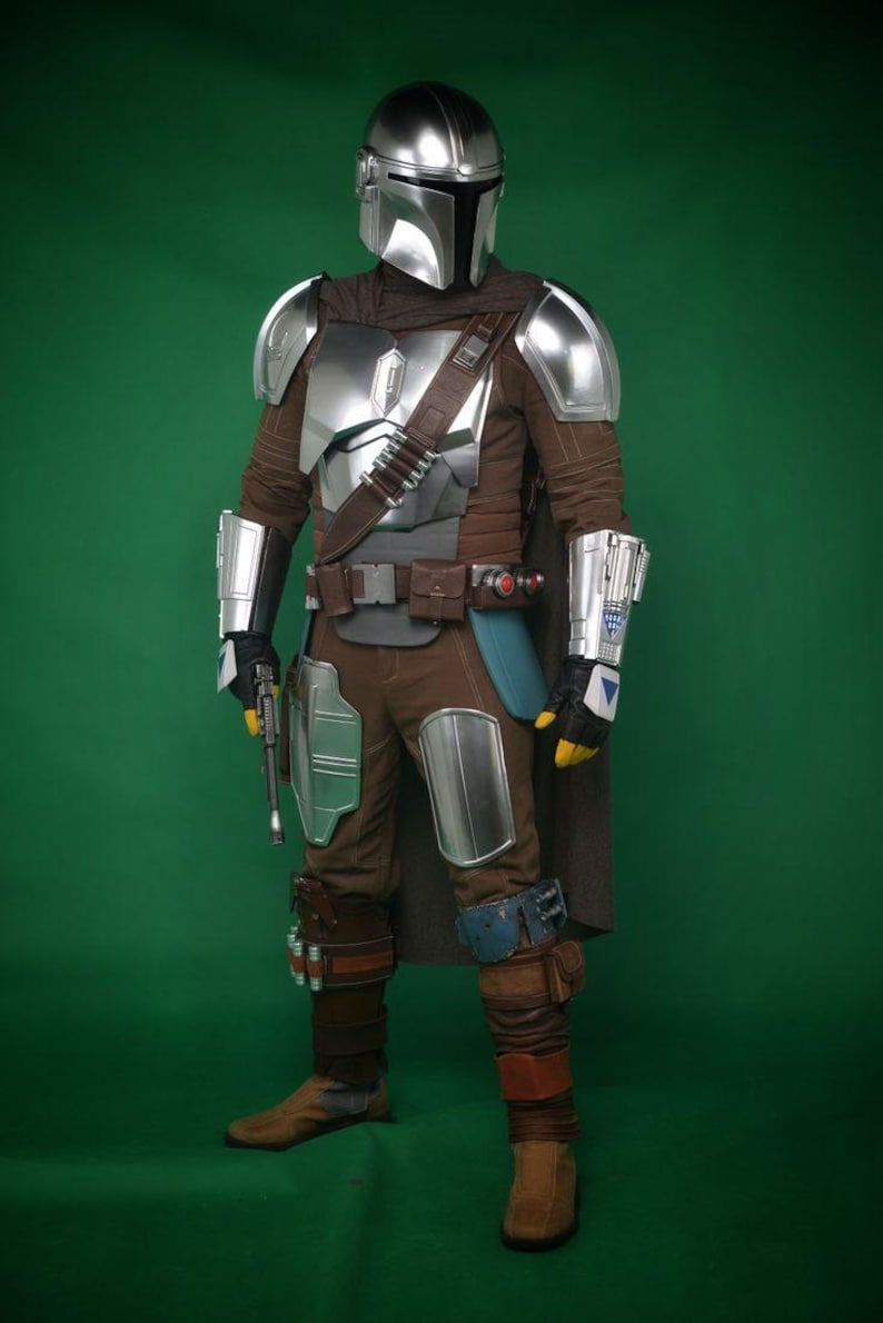 May include: A full-body shot of a Mandalorian costume. The costume features a silver helmet, chest armor, and arm guards. The outfit is brown with silver accents, a dark cape, and a utility belt. A blaster pistol is held in the left hand.