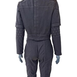May include: A dark gray jumpsuit with a cropped jacket and matching pants. The jacket features a high collar, shoulder accents, and geometric patterned detailing on the sleeves. White stitching outlines the seams. The jumpsuit is displayed on a mannequin.