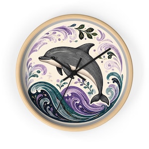 Könnte beinhalten: Eine runde Wanduhr mit hellem Holzrahmen zeigt in der Mitte einen grauen Delfin, der über stilisierte dunkel-türkisfarbene und violette Wellen springt. Der Hintergrund ist cremefarben mit dekorativen violetten Wirbeln und kleinen grünen Blättern.