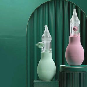 May include: Two nasal aspirators. One is light green, the other is pink. Both have a clear, conical tip and a bulb-shaped body. The aspirators are set against a dark green backdrop, with a matching green pedestal.