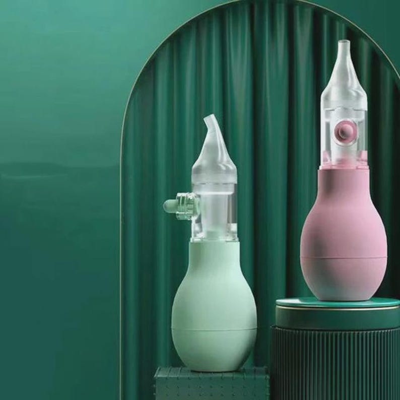 May include: Two nasal aspirators. One is light green, the other is pink. Both have a clear, conical tip and a bulb-shaped body. The aspirators are set against a dark green backdrop, with a matching green pedestal.