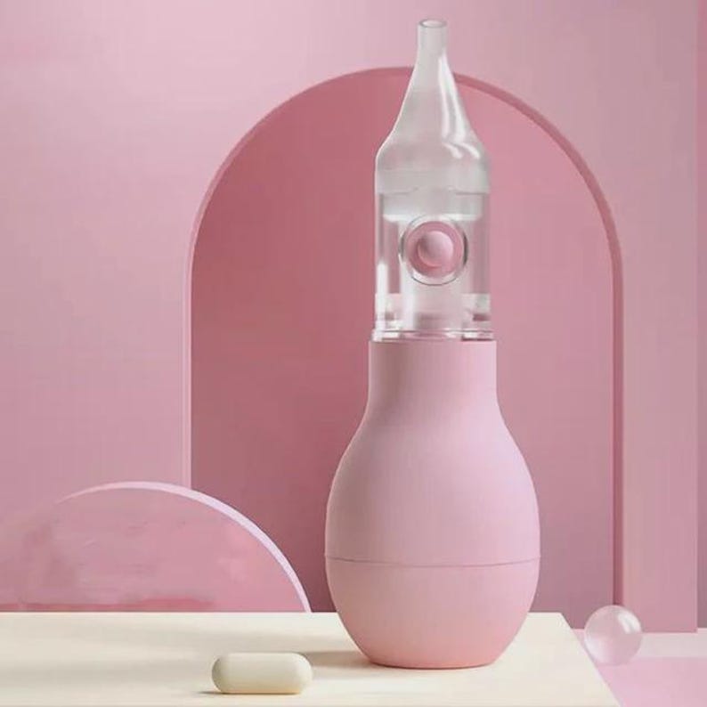 May include: A pink nasal aspirator with a clear, bottle-shaped top. The aspirator has a bulbous base and a clear, cylindrical section with a pink button. A small, white, oval object sits in front of the aspirator.