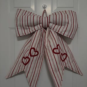 May include: A large, decorative bow made of white fabric with red vertical stripes. The bow features three embroidered red hearts on each of the long tails. The bow is hanging on a white door, perfect for Valentine's Day or other celebrations.