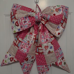 May include: A large, quilted bow in shades of pink, red, and white, with a patchwork design featuring hearts and other decorative elements. The bow is hanging on a white door, perfect for Valentine's Day or other special occasions.