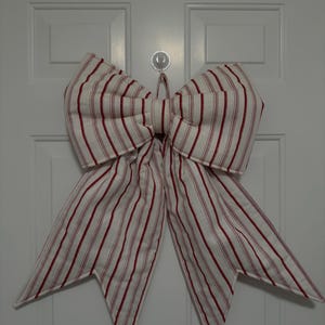 May include: A large, decorative bow made of white fabric with vertical red stripes. The bow is attached to a white door. The bow's design features a classic shape with multiple loops and long, pointed tails, suitable for holiday or seasonal decor.