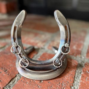 May include: A silver horseshoe with three small, round, silver rings attached. The horseshoe is displayed upright on a red brick surface. The horseshoe is a decorative item.