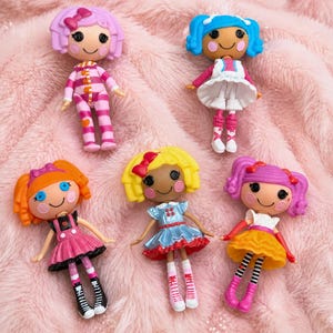 May include: Six small, colourful dolls with button eyes and rosy cheeks are arranged on a pink, textured surface. Each doll has a unique hairstyle and outfit, including dresses, striped leggings, and bows. The dolls are designed for children.