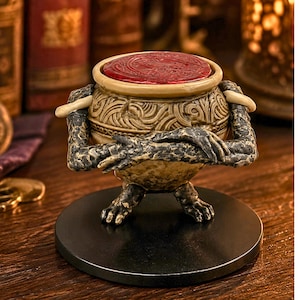 Peut inclure: Figurine détaillée d'un pot avec des bras et des jambes, sur une base noire. Le pot est beige avec des gravures complexes et un dessus circulaire rouge. Les bras sont croisés, et les mains sont détaillées.