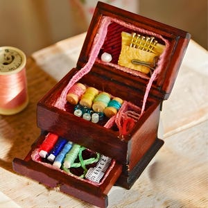 Puede incluir: Una pequeña caja de costura de madera abierta, llena de carretes de hilo de varios colores, agujas, alfileres y una cinta métrica. La caja está teñida de color marrón oscuro y forrada con tela rosa. Un cajón está abierto.