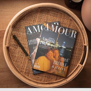 May include: Two magazines with the word "AMOUR" on the cover are on a woven tray. One magazine has the text "LOVERS & BEST FRIENDS" and the other says "since August 2021". A black pen rests on the tray.