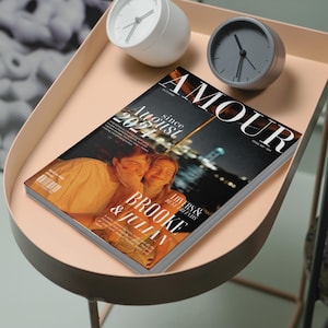 May include: A magazine titled "AMOUR" with the text "August 2024" and the names "Brooke & Julian" is displayed on a light pink tray table. Two clocks, one white and one gray, are also on the table. The magazine cover features a photo of two people.