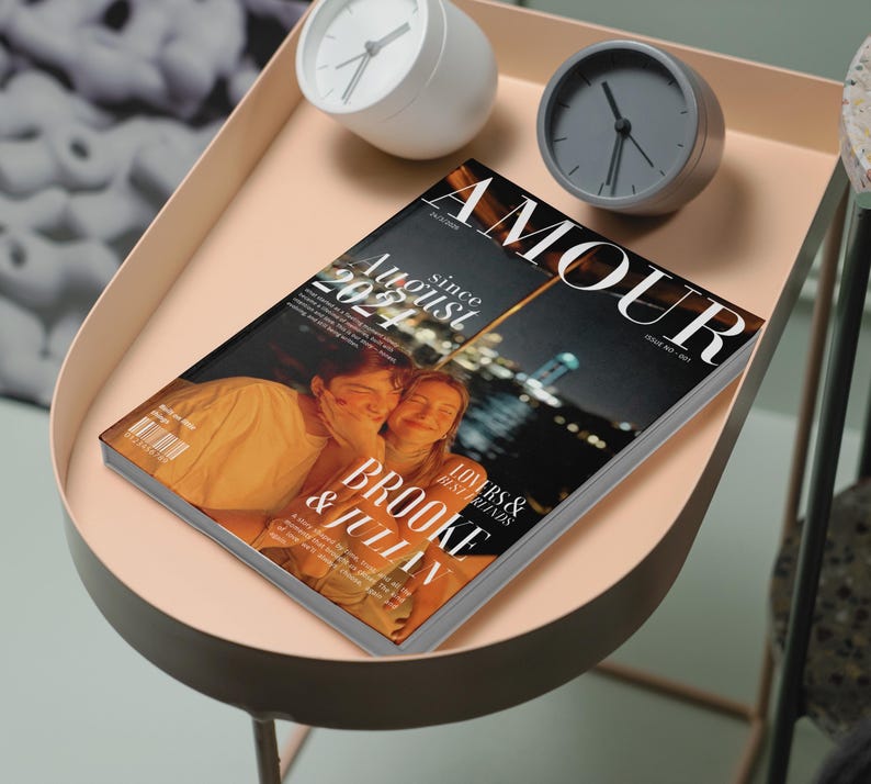 May include: A magazine titled "AMOUR" with the text "August 2024" and the names "Brooke & Julian" is displayed on a light pink tray table. Two clocks, one white and one gray, are also on the table. The magazine cover features a photo of two people.