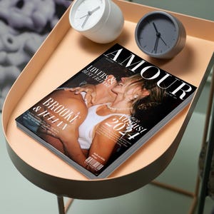 May include: A magazine titled "AMOUR" with a cover photo of a couple kissing. The magazine is on a peach-colored tray table, along with a white and a gray analog clock. The magazine cover reads "Brooke & Julian" and "Since August 2021."
