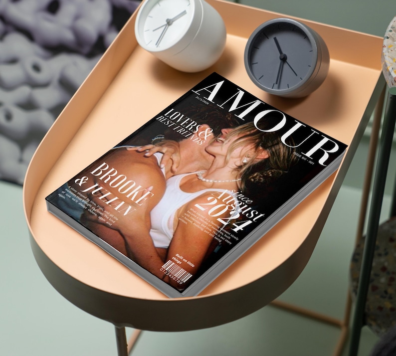May include: A magazine titled "AMOUR" with a cover photo of a couple kissing. The magazine is on a peach-colored tray table, along with a white and a gray analog clock. The magazine cover reads "Brooke & Julian" and "Since August 2021."