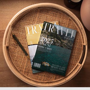 Può includere: Un vassoio rotondo in rattan intrecciato contiene due riviste e una penna. La copertina della rivista in alto recita "TRAVEL MAGAZINE" con "2025 AGOSTO" e "Brooke & Robin" stampati sotto. Una penna nera è appoggiata sul vassoio, accanto alle riviste.