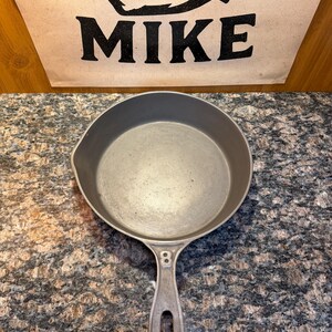 May include: A cast iron skillet with a long handle and a rounded cooking surface. The number "8" is stamped on the handle. The word "MIKE" is printed in black on a tan background.