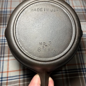 May include: A cast iron skillet with a dark, textured surface. The bottom of the pan is visible, displaying the text "MADE IN USA", "NO. 3", and "6 5/8 IN.". The handle extends from the side.