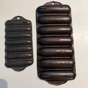 May include: Two antique cast iron Griswold stick pans. The larger pan is marked "No. 273" and the smaller pan is marked "No. 625". Both pans have the words "Crispy", "Corn", and "Stick Pan" embossed on them. The pans are dark brown.