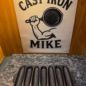 May include: A cast iron corn stick pan with seven corn-shaped molds. Above the pan is a sign with the words "CAST IRON MIKE" and an illustration of a muscular arm holding a skillet. The pan is dark brown.