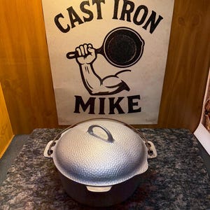 May include: A cast iron pot with a textured lid and a handle. The pot sits on a speckled surface, with a sign in the background that reads "CAST IRON MIKE" and features a graphic of a muscular arm holding a skillet.