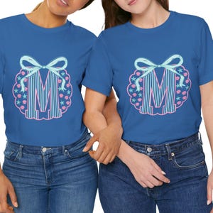 May include: Two blue t-shirts with a large letter "M" design. The design features a light blue bow, vertical stripes, and a border of pink seashells and starfish. The shirts are paired with blue jeans.