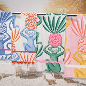 Pode incluir: Quatro toalhas de praia coloridas com desenhos de macacos e abacaxis. As toalhas apresentam macacos azuis, laranja, verdes e rosa segurando abacaxis com detalhes de estrelas. Estão penduradas em uma barra de metal, com uma cena de praia ao fundo.