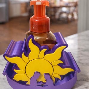 May include: A decorative soap dispenser holder in purple and yellow. The holder features a sun design with a silhouette of two figures. The dispenser has a brown bottle and an orange pump.